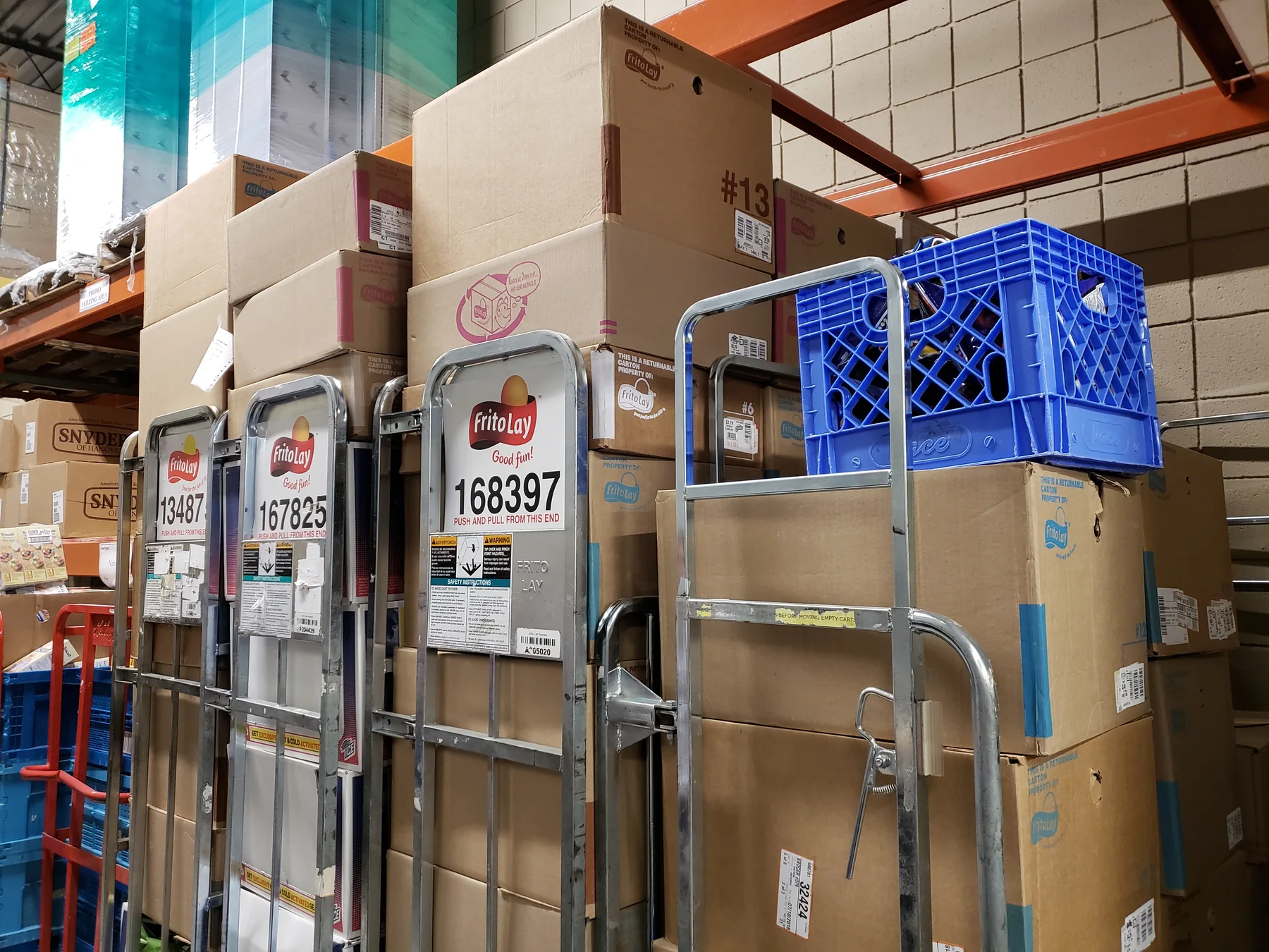 Frito-Lay delivery carts with large printed identification numbers, parked against racked stock in a warehouse aisle.